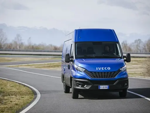 Niebieski Iveco Daily 3 na zakręcie drogi, z kierowcą za kierownicą. W tle góry i drzewa.