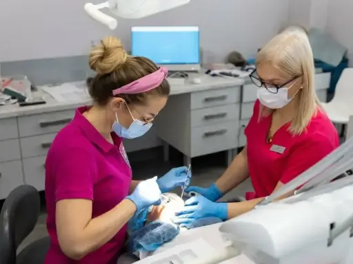 Dwie dentystki w maseczkach i rękawiczkach pracują przy pacjencie. Jedna z nich używa narzędzi, druga asystuje. Zabieg bez bólu dzięki lidokainie.