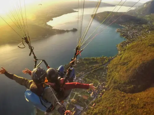 Vol en parapente biplace au-dessus d'un lac, avec un paysage de montagne et de village. Philippe Jeantot, malgré un accident passé, continue de voler.