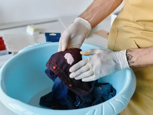 Hände mit Handschuhen behandeln ein dunkles Hemd mit einem hellen Fleck, um unbekannte Flecken zu entfernen.
