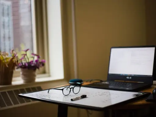 Okulary i pisak leżą na tablicy z zapiskami, obok laptopa. W tle okno z kwiatami. ibrus.