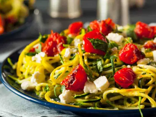 Pasta con verdure: spaghetti di zucchine, pomodorini confit, feta e basilico fresco su un piatto blu.