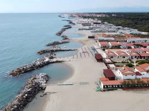 Vacker vy över pisa strand med sina stenpirar som bryter vågorna. Längre bort syns hus och en grönskande skog.