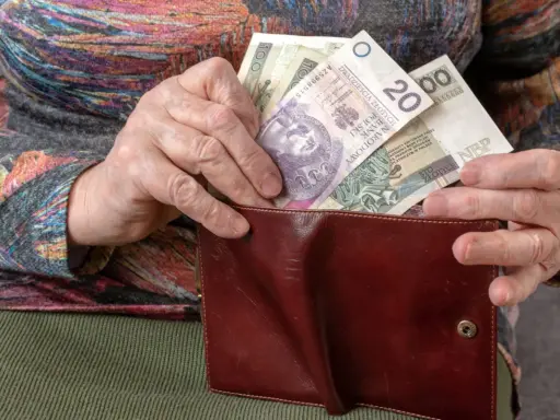 Dłoń starszej osoby wkłada polskie banknoty do brązowego portfela. Widać nominały 20, 100 i 50 złotych, stanowiące ekwiwalent codziennych wydatków.
