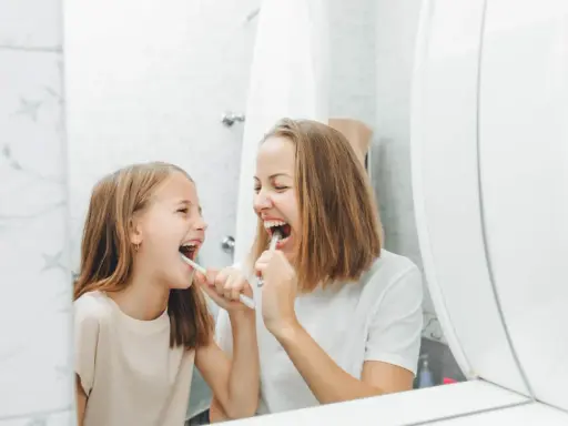 Mama i córka myją zęby, śmiejąc się. Dziecko nie chce myć zębów, ale mama przekonuje je zabawą.