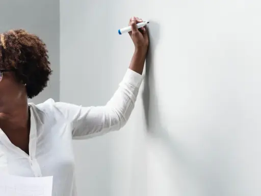 Frau mit Brille und Lockenwicklern hält einen Stift und bietet Nachhilfe an, während sie auf eine weiße Wand blickt.