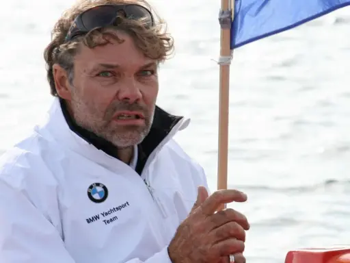 Markus Wieser, mit zerzaustem Haar und Sonnenbrille auf dem Kopf, trägt eine weiße BMW Yachtsport Team Jacke. Er blickt konzentriert auf das Wasser.