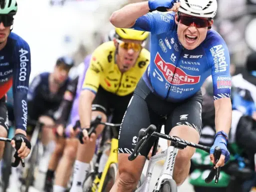 Radfahrer im blauen Trikot von Alpecin-Deceuninck jubelt nach einem Sieg, während andere Fahrer im Peloton folgen.