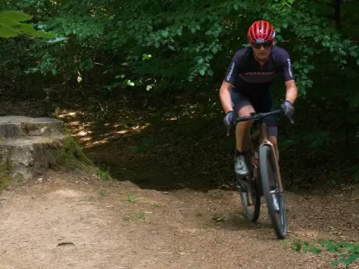 Radfahrer auf einem gravel dm-Pfad im Wald, mit einem Baumstumpf im Vordergrund.
