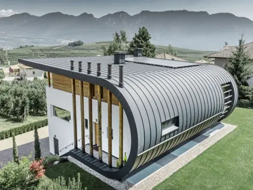 Nowoczesny dom z zaokrąglonym dachem z aluminium, które nie koroduje, otoczony zielenią i górami.