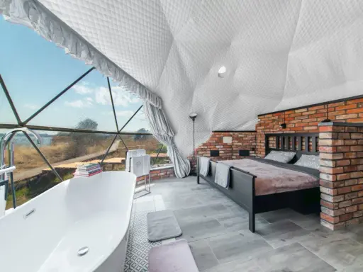 Luksusowy glamping nad jeziorem: wanna z widokiem, łóżko i ceglana ściana w kopułowym wnętrzu.