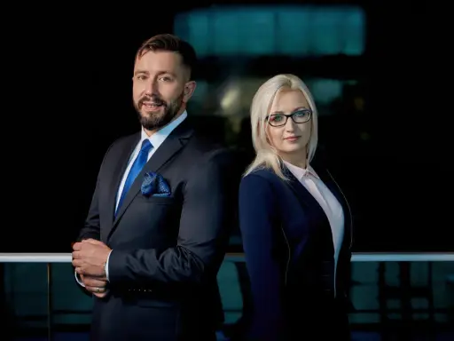 Para prawników w eleganckich garniturach, mężczyzna i kobieta, stojący na tle ciemnego biura.