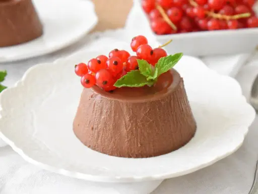 Panna cotta czekoladowa z czerwonymi porzeczkami i miętą na białym talerzyku.