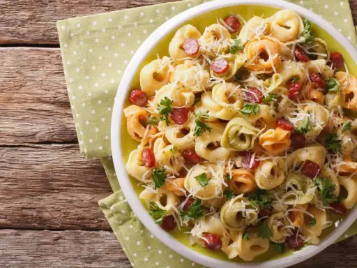 Kolorowa sałatka z tortellini, kawałkami kiełbasy i parmezanem, udekorowana natką pietruszki, podana na talerzu na zielonej serwetce w kropki.