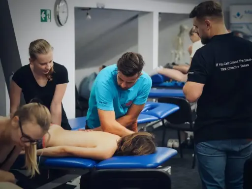 Uczestnicy szkolenia wykonują masaż tkanek miękkich na pacjencie leżącym na kozetce. Terapeuta w niebieskiej koszulce demonstruje technikę.