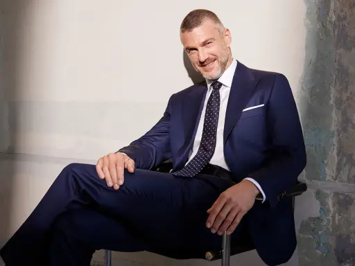 Uśmiechnięty mężczyzna w granatowym garniturze, z krawatem o klasycznej szerokości, siedzi w fotelu.