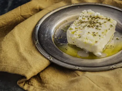 Ein Stück cremiger, vegetarischer Feta mit Kräutern und Olivenöl auf einem silbernen Teller, daneben ein rustikales Leinentuch.