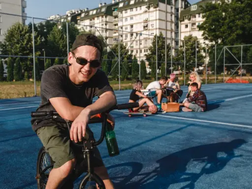 Uśmiechnięty chłopak na rowerze z butelką piwa, obok grupa znajomych. Może to być scena, która zakończy się mandatem za jazdę rowerem po alkoholu.