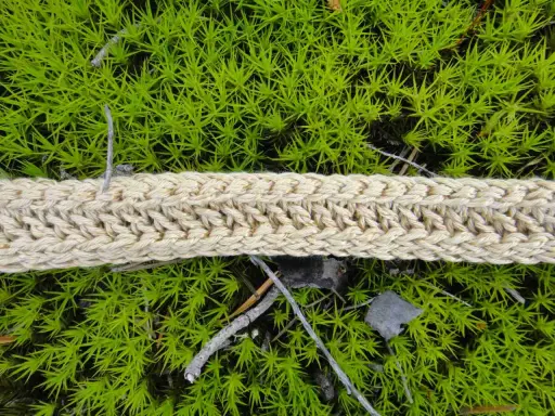 Ett naturfärgat virkat band ligger på grön mossa. Bandet har ett tydligt flätmönster.