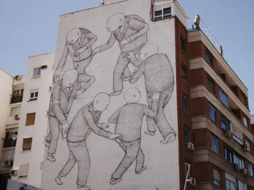 A striking street art style mural depicts a group of bald figures in suits, seemingly engaged in a peculiar, interconnected dance or struggle on a building's facade.