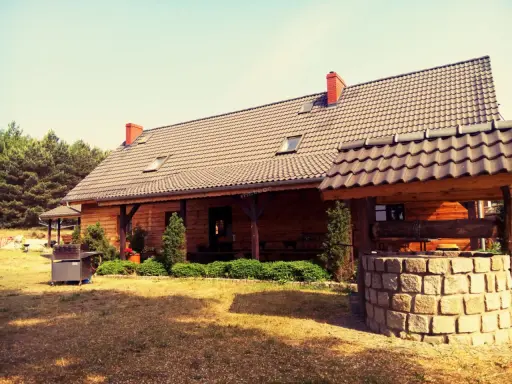 Drewniany dom pod kasztanem, z kamienną studnią i grillem, otoczony zielenią i lasem.