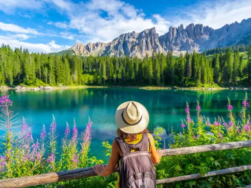 En kvinna med hatt och ryggsäck njuter av den vackra **Dolomiterna sommar**-utsikten över en turkos sjö, omgiven av skog och berg.