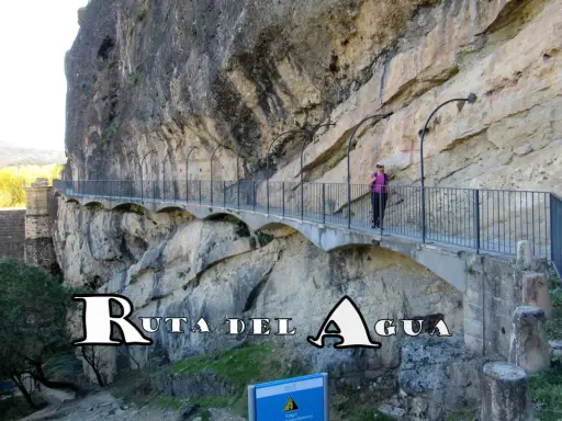 Un pontón de la oliva, parte de la Ruta del Agua, se aferra a un acantilado rocoso. Una mujer camina por el sendero con barandillas.