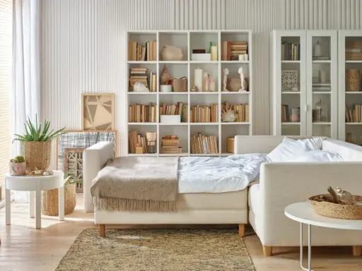 Jasny salon z sypialnią na małej powierzchni. Rozkładana sofa, regały z książkami i dekoracje tworzą przytulną przestrzeń.