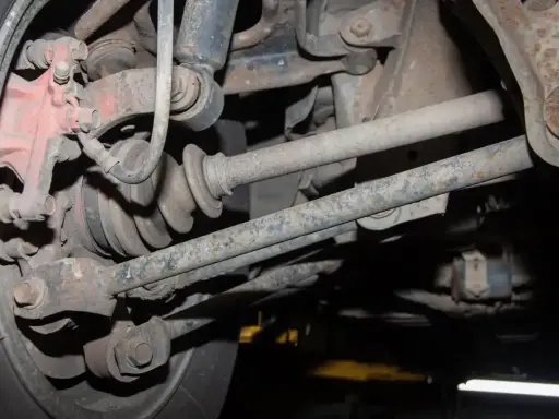 Widok od spodu na skorodowane elementy zawieszenia samochodu, w tym przegub napędowy i wahacze, tuż pod nadkolem.