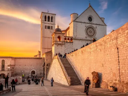 Assisi cosa vedere: la Basilica di San Francesco al tramonto, con la sua imponente facciata e la torre campanaria, illuminata da una calda luce dorata.