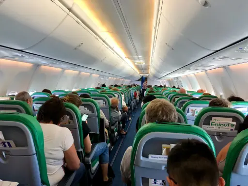 Wnętrze samolotu Transavia, pasażerowie siedzą w rzędach, przygotowani do lotu. Bagaż podręczny schowany w schowkach nad głowami.