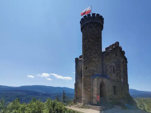 Kamienny zamek henryka na wzgórzu, z polską flagą na wieży, góruje nad zielonymi lasami i błękitnymi górami.