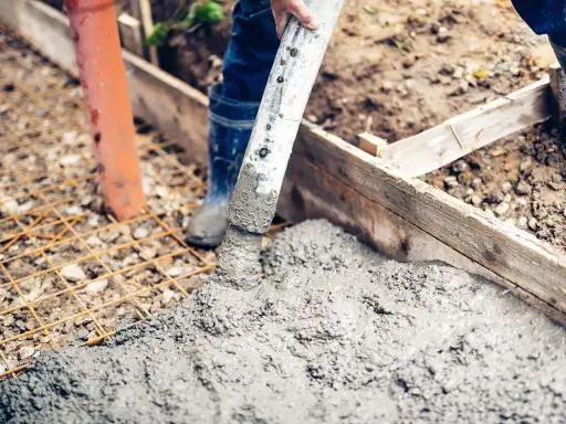 Arbeiter betonieren bei Regen eine Fundamentplatte. Beton fließt aus einem Schlauch auf eine Bewehrung.