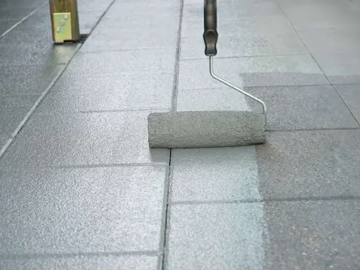 Ein Farbroller streicht graue Pflastersteine. Die rechte Seite ist frisch gestrichen, die linke Seite noch ungestrichen.