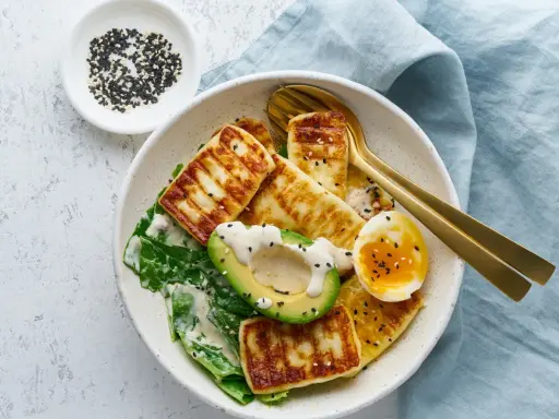 Szybki keto obiad: grillowany ser halloumi, awokado, jajko na miękko i zielone liście, posypane sezamem.