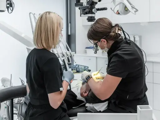 Dwie dentystki pracują przy pacjencie. Jedna używa mikroskopu, druga trzyma narzędzie. To obrazek pokazujący, jak zostać dentystą.