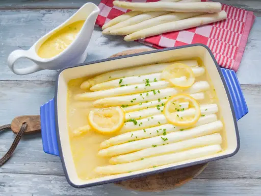 Weißer Spargel in einer Auflaufform mit Zitrone und Kräutern, daneben eine Sauciere mit Sauce.