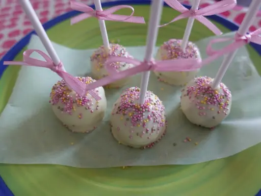 Sju söta cake pops med vit choklad och rosa, gula och lila strössel, dekorerade med rosa band.