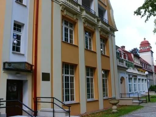 Sanatorium Jan Kazimierz w Dusznikach-Zdroju, z charakterystyczną żółto-kremową fasadą i czerwonym dachem, otoczone zielenią.