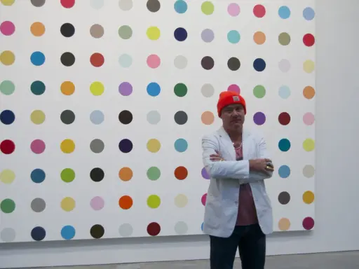Damien Hirst steht vor seiner berühmten Punkt-Malerei, gekleidet in eine orangefarbene Mütze und einen weißen Blazer.