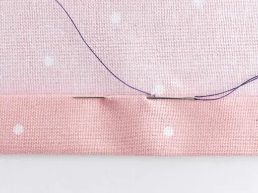 En nål med lila tråd sys genom rosa tyg med prickar. Stygnen är nästan osynliga, ett tecken på skickligt hantverk.