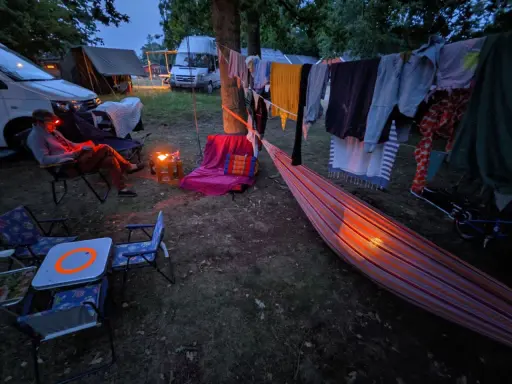 Una persona lee en una silla junto a una fogata, ropa secándose al aire y una hamaca iluminada. Disfrutando de una tranquila acampada al aire libre.