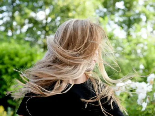 Warkocze wiatru rozwiewają chłodny blond włosów dziewczynki na tle zieleni.