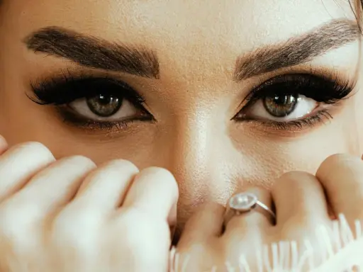 Yeux verts intenses, cils longs et maquillage sourcils parfait. Mains délicates ornées de bagues cachent une partie du visage.