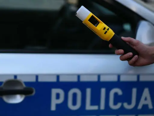 Policjant przeprowadza badanie trzeźwości nieletniego przy użyciu alkomatu. Na samochodzie widnieje napis "POLICJA".