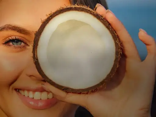 Donna sorride con un fiore tra i capelli, coprendo un occhio con una noce di cocco. Attenzione: olio di cocco sul viso fa male.