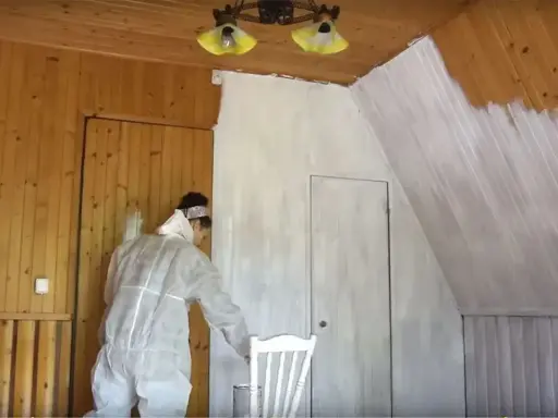 Osoba w kombinezonie maluje lakierowaną boazerię bez szlifowania na biało.