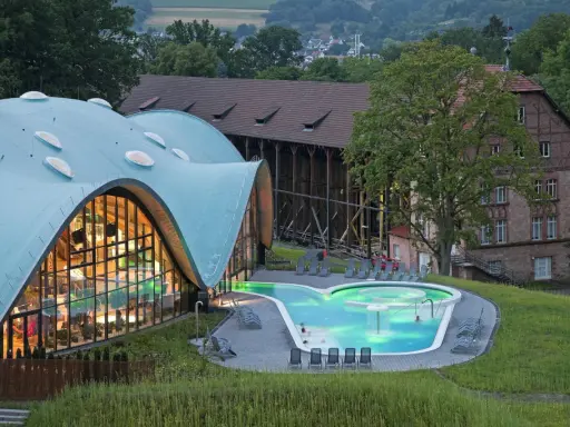 Moderne Therme mit geschwungenem Dach und beleuchtetem Pool, daneben historische Gebäude.