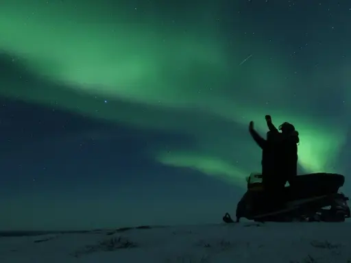 En person på snöskoter i Kiruna Lapland sträcker upp armarna mot norrskenet som lyser grönt på natthimlen.