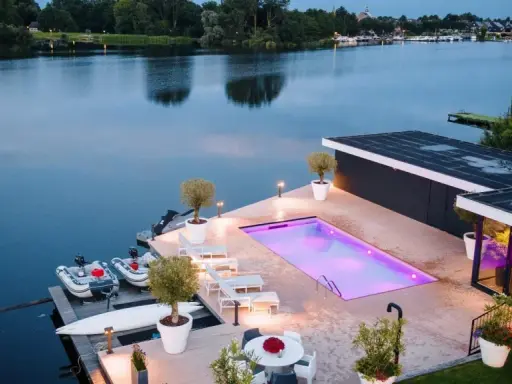 Lyxig pool med platssvetsad liner och belysning, omgiven av solstolar och växter. Båtar ligger vid bryggan.